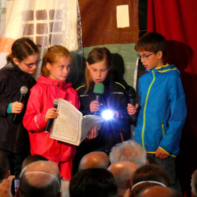 Im Bild sind vier Kinder mit Mikrofon und einer Taschenlampe sichtbar. Sie schauen in ein aufgeschlagenes Textheft, aus dem vorgelesen wird.