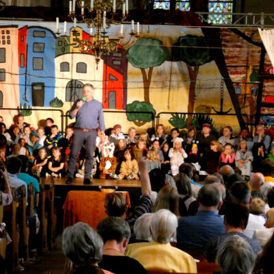 Im Bild ist die volle Kirche zu sehen. Auf der Bühne hocken Kinder in mehreren Reihen. Die Kirchenbänke sind voll besetzt. Ein Mann steht in Zentrum der Bühne und spricht ins Mikrofon.
