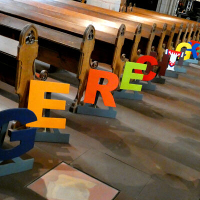 Im Bild sieht man die einzelnen Buchstaben des Wortes "Gerechtigkeit" zwischen den Kirchenbänken.