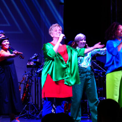 Im Bild steht eine Gruppe von singender Damen auf der Bühne. Im Hintergrund sind Musikinstrumente sichtbar.