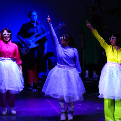 Im Bild sind drei Sängerinnen in Kostümen sichtbar.
