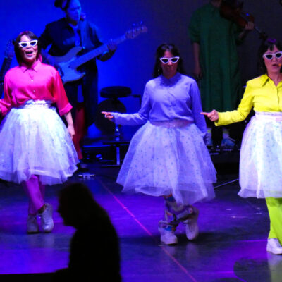 Im Bild sind drei Sängerinnen in Kostümen sichtbar.